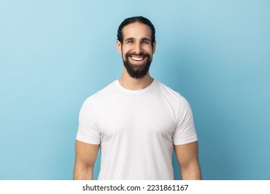 Portrait of bearded handsome man wearing white T-shirt standing looking at camera with satisfied face and smiling, expressing happiness. Indoor studio shot isolated on blue background. - Shutterstock ID 2231861167