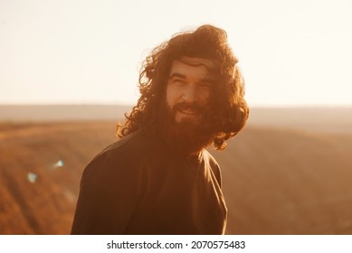 Portrait Of Bearded Hipster Man With Long Hair In Windy Day During Sunset On Hills