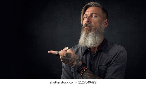 Portrait of a bearded hipster, who is pointing with his finger to the left side, isolated on a black background