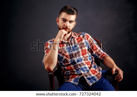 Portrait of bearded male with long hair sitting on chair and looking at camera over dark background in studio.