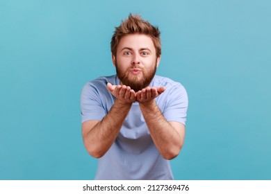 Portrait of bearded man blowing air kiss keeps lips, folded palm towards mouth, sends mwah coquettish, wants to cheer up her partner. Indoor studio shot isolated on blue background.