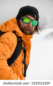 Portrait Of Bearded Man In Bright Jacket And Black Hat With Glasses With Reflection Of Winter Ski Resort.