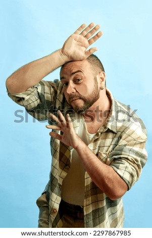 Portrait of bearded man in checkered shirt leaning on transparent glass against blue studio background. Concept of human emotions, lifestyle, facial expression, news, sales Ad