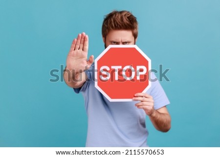 Portrait of bearded man covering half of face with octagonal Stop symbol, showing red traffic sign and ban gesture with palm, warning of forbidden way. Indoor studio shot isolated on blue background.