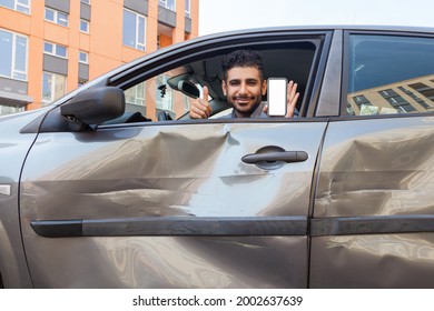 Portrait Of Bearded Man In Crashed Car Looking At Camera And Smiling, Showing Thumb Up And Smart Phone With Blank Screen, Advertising Insurance Inspection Services After Road Accident. Outdoor Shot.