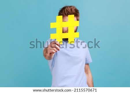 Portrait of bearded man hiding behind yellow hashtag symbol, covering face with hash sign, concept of blogging and viral topics on internet. Indoor studio shot isolated on blue background.