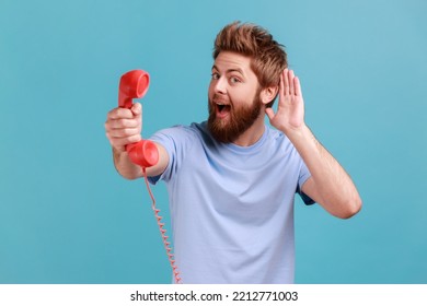 Portrait Of Bearded Man Holding And Showing Retro Phone Handset To Camera, Asking To Answer Phone, Keeps Hand Near Ear, Hearing Your Talking. Indoor Studio Shot Isolated On Blue Background.