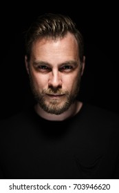 Portrait Of A Bearded Man With An Intense Look Standing In A Dark Room Against A Black Background.