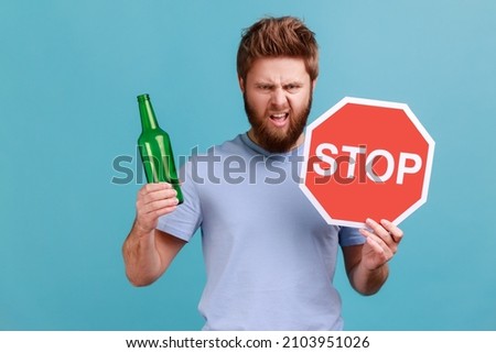 Portrait of bearded man showing alcoholic beverage beer bottle and stop sign, warning and worrying, looking at camera with angry expression. Indoor studio shot isolated on blue background.