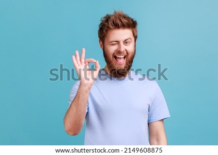 Portrait of bearded man showing okay sign, success sign, satisfied with good service, looking at camera and winking, expressing positive emotions. Indoor studio shot isolated on blue background.