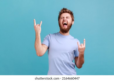 Portrait of bearded man showing rock and roll gesture heavy metal sign, enjoys favorite music on party, has fun squints face exclaims from joy. Indoor studio shot isolated on blue background. - Shutterstock ID 2164660447