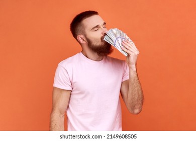 Portrait Of Bearded Man Smelling Dollar Banknotes With Pleasure Expression, Enjoying Big Money Profit And Rich Life, Lottery Win, Wearing Pink T-shirt. Indoor Studio Shot Isolated On Orange Background