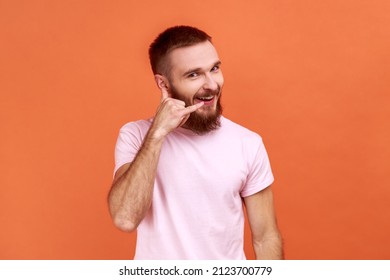 Portrait Of Bearded Man Standing With Telephone Hand Gesture And Smiling To Camera, Flirting Offering To Contact By Phone, Wearing Pink T-shirt. Indoor Studio Shot Isolated On Orange Background.