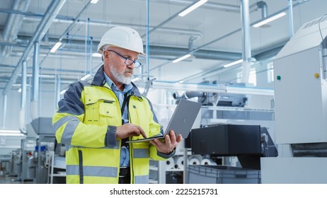 Portrait of a Bearded Middle Aged Engineer Standing in a Factory Facility, Wearing a High Visibility Jacket and a White Hard Hat. Heavy Industry Specialist Working on Laptop Computer. - Powered by Shutterstock