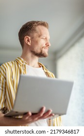 Portrait Of Bearded Middle Aged Man Holding Open Laptop Computer, Looking Away And Thinking Standing Indoors. Deep In Thought Male Working Online From Home Or Browsing Internet On Pc