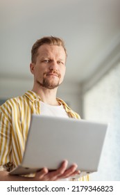 Portrait Of Bearded Middle Aged Man Holding Open Laptop Computer And Looking At Camera Standing Indoors. Male Working Online From Home Or Browsing Internet On Pc