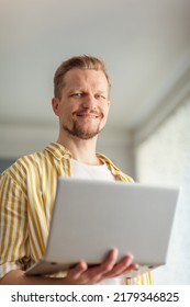 Portrait Of Bearded Middle Aged Man Holding Open Laptop Computer, Looking At Camera And Smiling Cheerfully Standing Indoors. Male Working Or Studying Online From Home