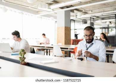 Portrait Of Bearded Middle-Eastern Businessman Drinking Coffee At Work And Watching Videos From Smartphone In Open Office, Copy Space