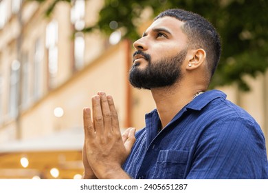 Portrait of bearded religion indian man praying with closed eyes to God asking for blessing, help, forgiveness outdoor. Adult guy clasping hands wishing luck in urban sunny city street. Town lifestyle