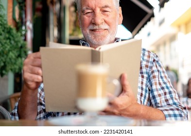Portrait of bearded senior man sitting outdoor at a cafe table reading a book while enjoying a coffee and milk drink - caucasian elderly man relaxed in retirement or vacation - Shutterstock ID 2231420141