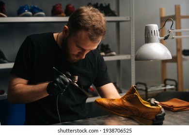 Portrait of bearded shoemaker wearing black gloves spraying paint of light brown leather shoes, close-up. Concept of cobbler artisan repairing and restoration work in shoe repair shop. - Powered by Shutterstock