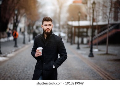 Portrait Of A Bearded Stylish Young Handsome Man In Black Coat Is Drinking Coffee Outside. 
