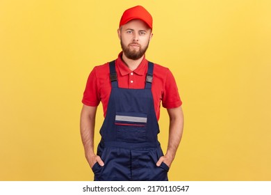 Portrait Of Bearded Worker Man Wearing Blue Uniform And Red Cap Standing With Hands In Pockets, Looking At Camera With Serious Facial Expression. Indoor Studio Shot Isolated On Yellow Background.