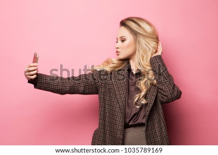 Portrait of beautifu  smiling blond woman in casual clothes with perfect  makeup with smartphone, makes selfie over pink background