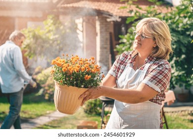 Portrait of a beautiful adult woman holding a flowerpot in the backyard. Senior couple cleaning their garden during the sunny day. - Powered by Shutterstock