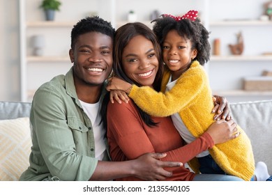 Portrait Of Beautiful African American Family Of Three Embracing At Home, Cheerful Young Black Parents And Little Daughter Hugging And Smiling At Camera, Enjoying Spending Time Together, Closeup