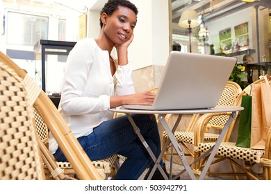 Portrait Of Beautiful African American Professional Woman Sitting In Coffee Shop Using A Laptop Computer, Working Outdoors. Smart Black Business Woman In City, Thoughtfully Smiling In Exterior.