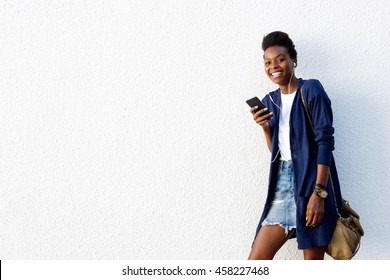 Portrait Of Beautiful African American Woman Standing Against White Wall And Listening To Music On Mobile Phone 
