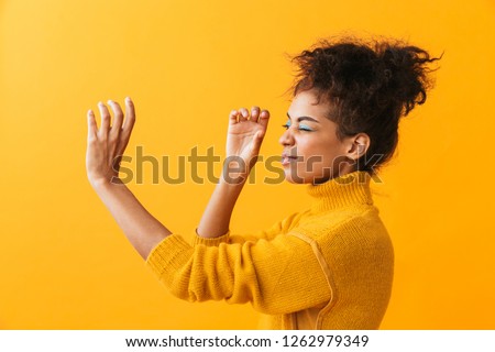 Portrait of beautiful african american woman with afro hairstyle looking through invisible spyglass isolated over yellow background