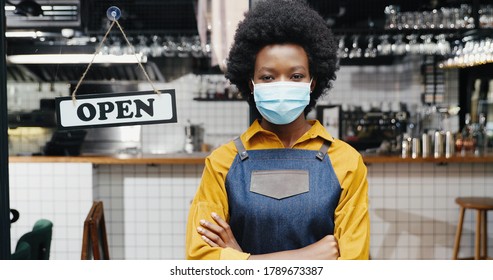 Portrait of beautiful African American young woman waitress in apron and medical looking to camera with table open at cafe entrance outdoor. Pretty barrista standing at bar door with board open Reopen - Powered by Shutterstock