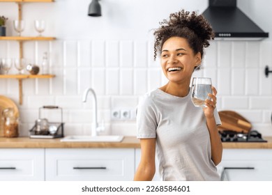 Portrait of a beautiful African American young woman drinking water standing in the kitchen at home looking away and smiling, wellness healthy food concept - Powered by Shutterstock