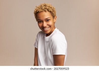 Portrait of beautiful African woman with big toothy smile, looking at camera during studio shooting, dressed in white unisex T-shirt, having nose and ears piercing on clear young face with no makeup