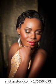 Portrait Of Beautiful African Woman Dressed As A Jazz Age Diva With Gold Makeup, Red Lips And Diamond Earrings, Posing In Front Of A Black Background.