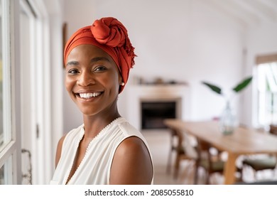 Portrait of a beautiful african woman smiling while looking at camera. Mid adult woman with traditional african headscarf stay at home and smiling. Cheerful mature lady standing near the window. - Shutterstock ID 2085055810