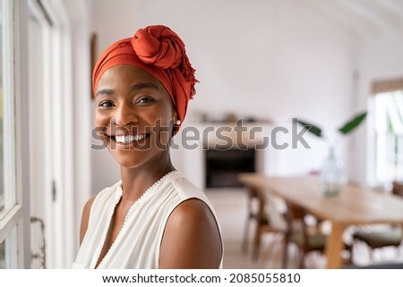 Photo of Portrait of a beautiful african woman smiling while looking at camera. Mid adult woman with traditional african headscarf stay at home and smiling. Cheerful mature lady standing near the window.