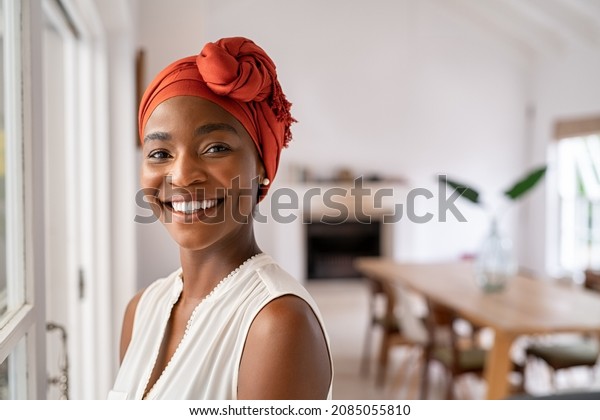 Portrait of a beautiful african woman smiling\
while looking at camera. Mid adult woman with traditional african\
headscarf stay at home and smiling. Cheerful mature lady standing\
near the window.