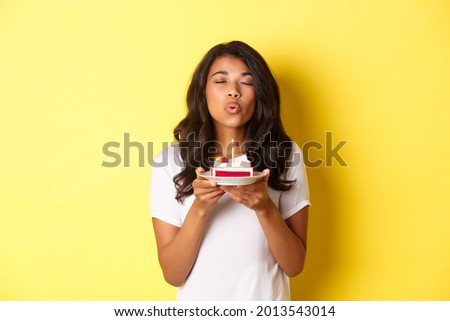 Portrait of beautiful african-american girl celebrating birthday, blowing lit candle on b-day cake and making a wish, standing over yellow background