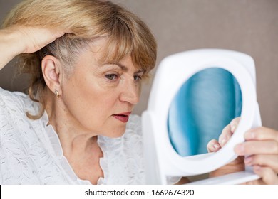 Portrait of a beautiful aged woman in glasses at home looking in the mirror. Beautiful senior woman checking gray hair. The concept of the aging process.