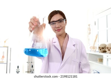 Portrait Of Beautiful Asian Female Scientist Wear Lab Coats And Safety Glasses, Happy Smiling Researcher Woman Standing And Showing Blue Chemical Flask In Laboratory, Woman Work  In Science Field.