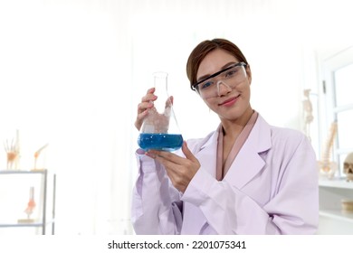 Portrait Of Beautiful Asian Female Scientist Wear Lab Coats And Safety Glasses, Happy Smiling Researcher Woman Standing And Showing Blue Chemical Flask In Laboratory, Woman Work  In Science Field.