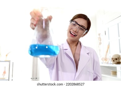 Portrait Of Beautiful Asian Female Scientist Wear Lab Coats And Safety Glasses, Happy Smiling Researcher Woman Standing And Showing Blue Chemical Flask In Laboratory, Woman Work  In Science Field.