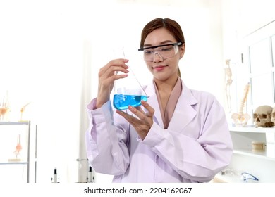 Portrait Of Beautiful Asian Female Scientist Wear Lab Coats And Safety Glasses, Happy Smiling Researcher Woman Standing And Showing Blue Chemical Flask In Laboratory, Woman Work  In Science Field.