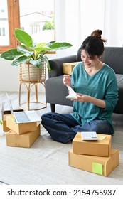 Portrait, Beautiful Asian Millennial Woman Business Startup Entrepreneur Notes Something On A Spiral Notepad While Preparing The Delivery Boxes.