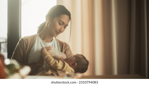 Portrait of Beautiful Asian Mother Feeding Milk to her Infant Using a Baby Bottle at Bright Home. Young Woman New to Motherhood Bonding with her Child and Enjoying an Affectionate Family Moment.