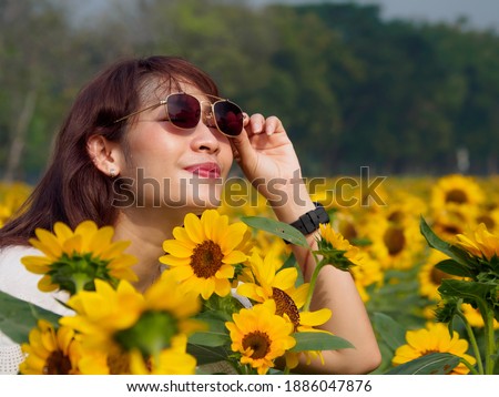 Portrait of a beautiful Asian woman around 30-40 years old inwhite shirt, cream hat, and black glasses. Standing in field yellow sunflowers blooming onday relaxation. With pleasure from  work