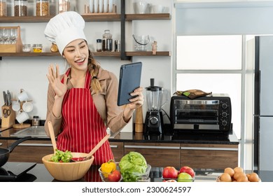 Portrait of beautiful Asian woman in chef's hat and apron use digital tablet video call while cooking in kitchen. Pan and electric stove, eggs and equipment on the counter. copy space - Powered by Shutterstock